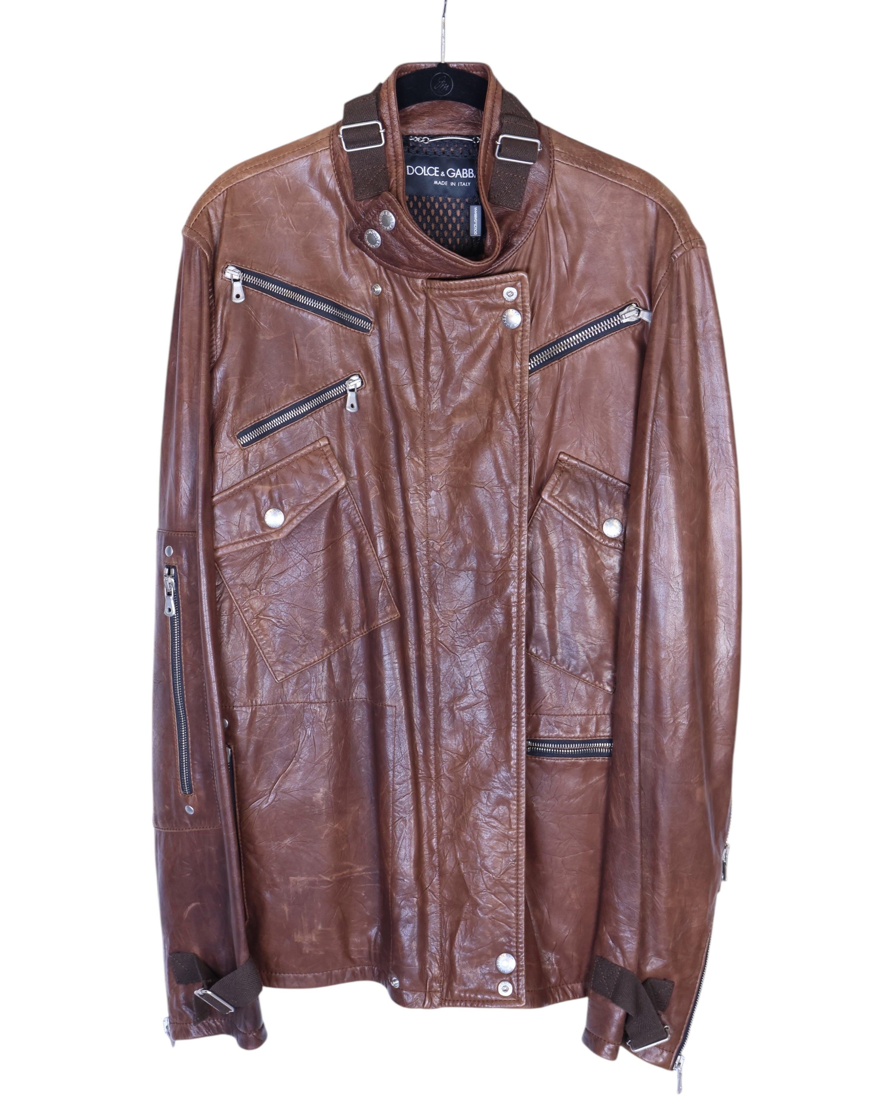 A brown Dolce & Gabbana sheepskin leather jacket with multiple zippers and a collar. Dolce & Gabbana brown/cognac sheepskin leather biker jacket; asymmetrical snap-over front, funnel collar with throat latch, multiple zip and snap-flap pockets, zip cuffs, buckle straps at hem; silver-tone hardware; regular fit, unisex; great with denim, tees, boots; moto, biker, rocker, Y2K; brand context: Italian luxury house, made in Italy, artisanal finish; vintage, lightly distressed texture.