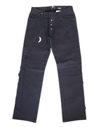 A pair of black jeans with shark teeth pendants displayed against a white background. Vintage Voyage Mazzilli Michielsens black jeans/denim trousers with straight-leg cut, visible button fly and five pockets, plus tonal stitched/appliqué trims along the seams. Finished with a waist drawcord carrying a shark-tooth–style pendant charm and back grommet lacing; luxury, rare and unique, laid flat on white.