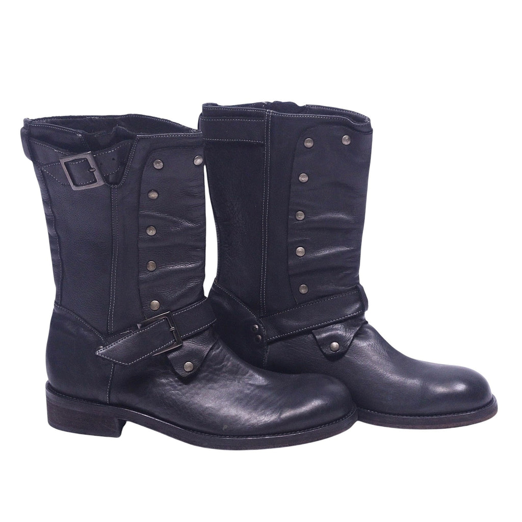 A pair of John Varvatos black leather boots with a buckled strap and metal studs on the side. John Varvatos black leather mid-calf boots with round toe, stacked heel and harness-style buckle strap, plus studded front panels and a top buckle. Grained finish with a rugged moto/engineer vibe; vintage, designer, rare.