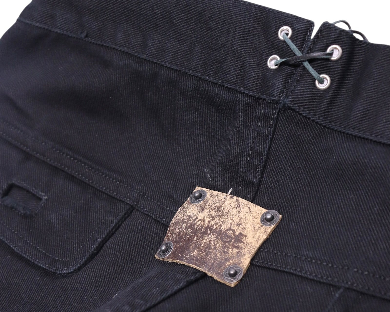 Back-waist detail of black Voyage Mazzilli Michielsens jeans featuring grommet lacing and a distressed square leather logo patch riveted to the waistband, with clean tonal stitching on twill denim.