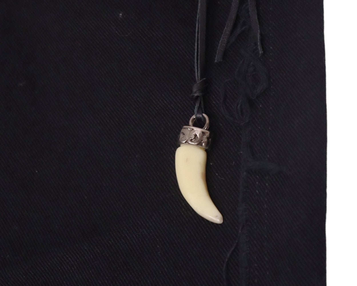 Close-up of a shark-tooth–style pendant charm with metal cap hanging from a leather cord at the waistband of black Voyage Mazzilli Michielsens jeans, showing tonal stitched/appliqué seam detail on twill denim.