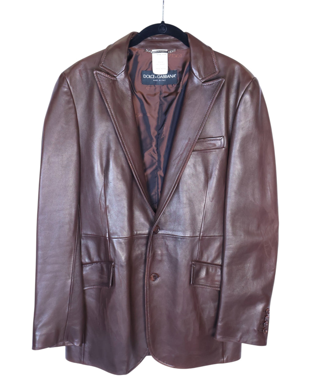 A brown leather jacket with long sleeves, collar, and front pockets, displayed on a white background. Dolce & Gabbana chocolate/brown leather blazer jacket; supple calfskin, notch lapels, single-breasted two-button front, chest welt pocket + flap pockets, buttoned cuffs, full logo lining; modern tailored fit, unisex; pairs with denim, tailoring, boots; style tags: luxe, minimal, 90s/Y2K; brand context: Italian luxury house, artisanal Made in Italy.