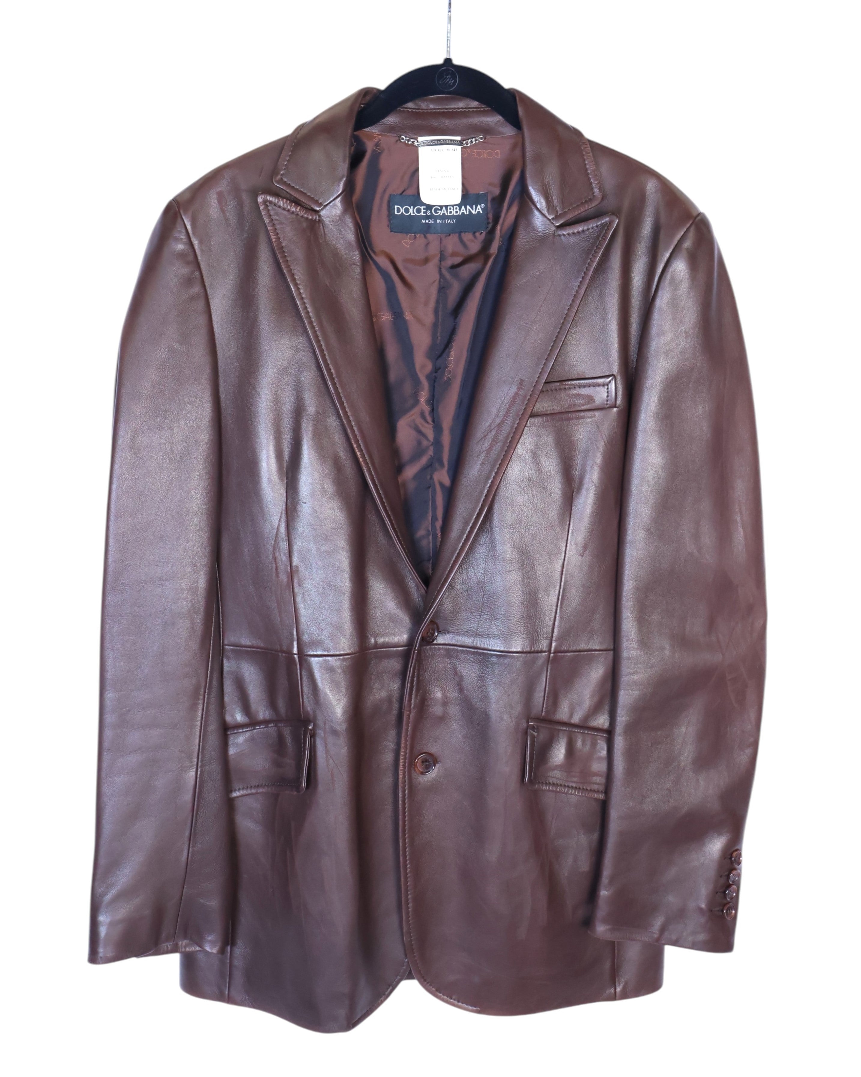 A brown leather jacket with long sleeves, collar, and front pockets, displayed on a white background. Dolce & Gabbana chocolate/brown leather blazer jacket; supple calfskin, notch lapels, single-breasted two-button front, chest welt pocket + flap pockets, buttoned cuffs, full logo lining; modern tailored fit, unisex; pairs with denim, tailoring, boots; style tags: luxe, minimal, 90s/Y2K; brand context: Italian luxury house, artisanal Made in Italy.