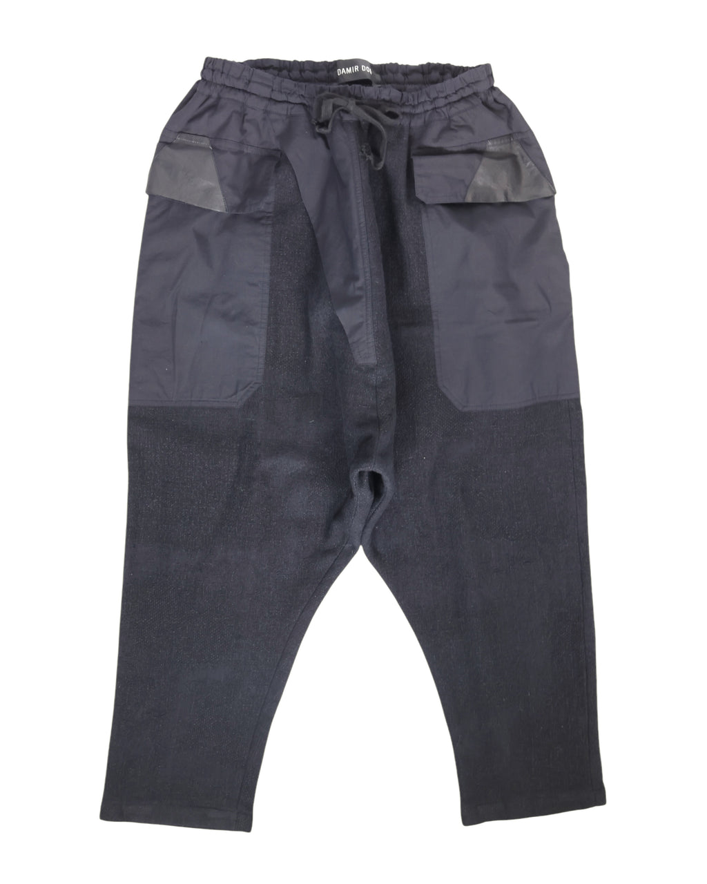 Second-hand vintage Damir Doma black sweatpants/joggers with drawstring waist and drop-crotch, tapered cropped legs, oversized front patch pockets with leather-trimmed flaps and mixed-fabric panels; minimalist avant-garde streetwear, rare designer piece laid flat on white.