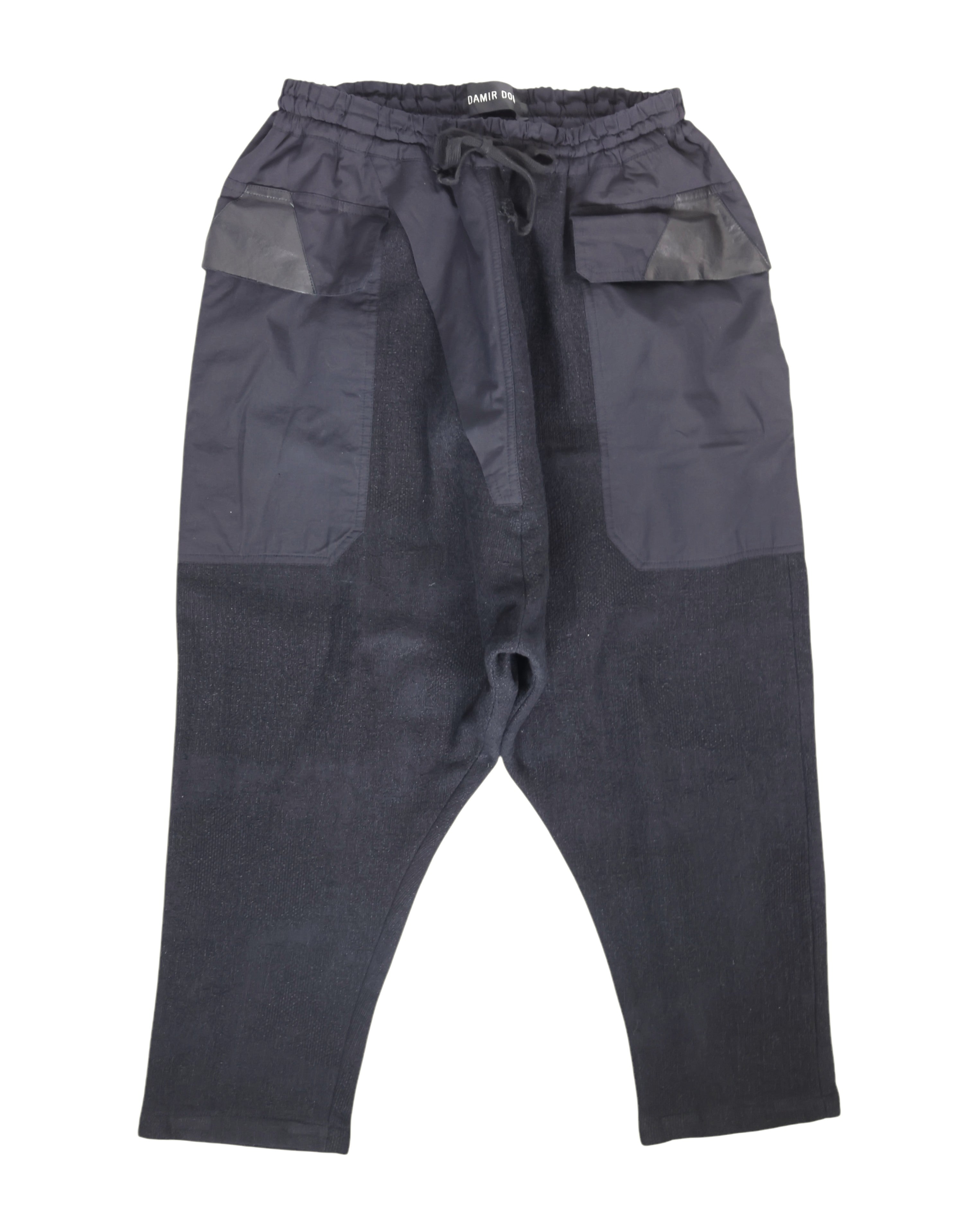 Second-hand vintage Damir Doma black sweatpants/joggers with drawstring waist and drop-crotch, tapered cropped legs, oversized front patch pockets with leather-trimmed flaps and mixed-fabric panels; minimalist avant-garde streetwear, rare designer piece laid flat on white.