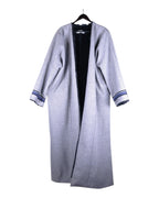 A long, gray faux fur lined jacket with blue accents on the cuffs, displayed on a hanger. Abadia grey farwa coat/jacket/overcoat/duster/robe; open-front maxi length, dropped shoulders, wide sleeves with striped cuffs; plush faux-fur lining for warmth; oversized relaxed fit, unisex-friendly; great over abayas, dresses, denim, for travel and winter layering; tags: modest, minimalist, bohemian, contemporary, heritage; Abadia (Saudi, artisan-led, Middle-Eastern craft roots), limited piece.