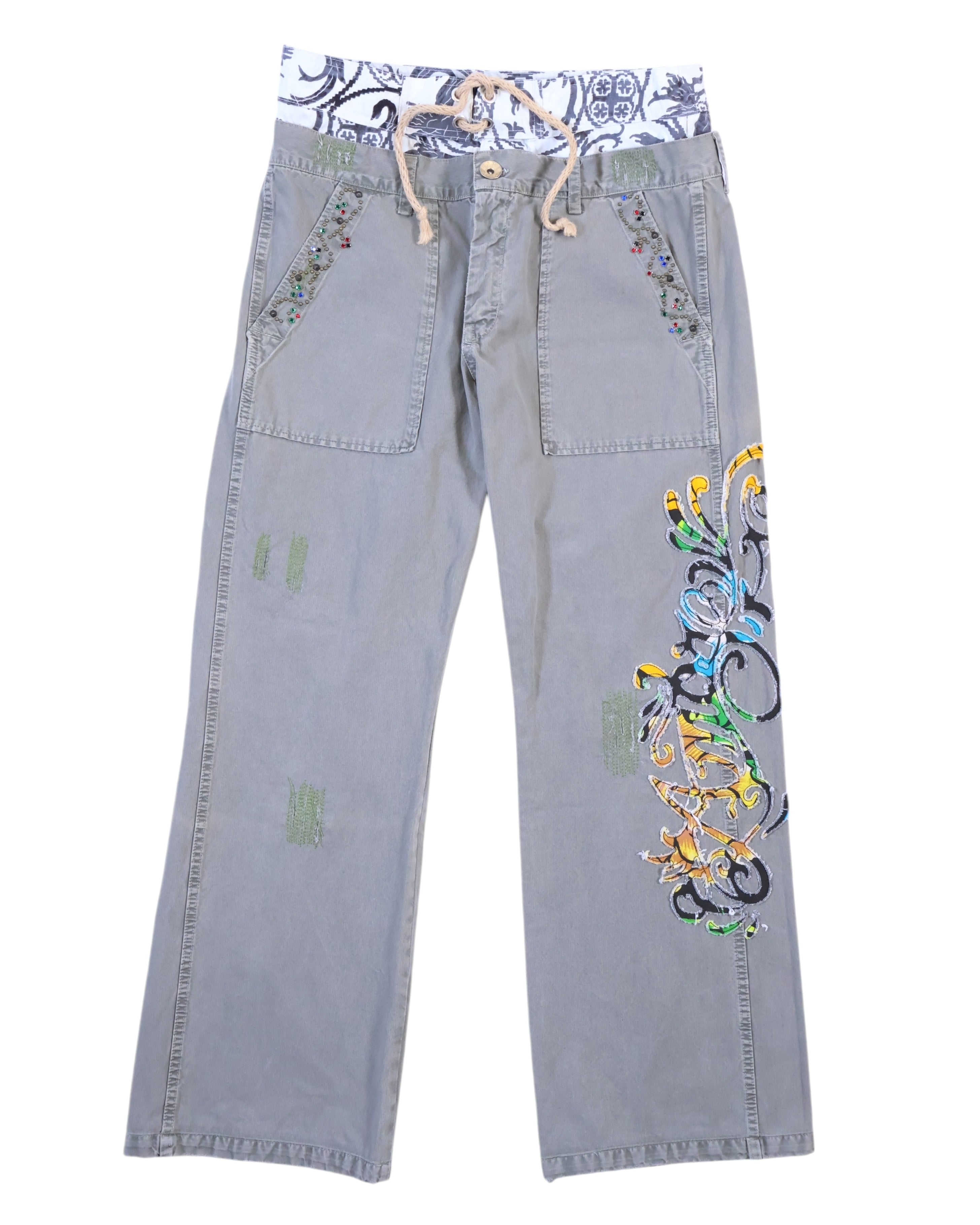 RA-RE washed grey trousers with relaxed straight legs, large front patch pockets with studded trim, visible repair patches, and a bold multicolour scroll embroidery down the right leg. A contrast printed waistband with rope drawcord peeks out; boho-meets-streetwear, designer, second-hand and unique.