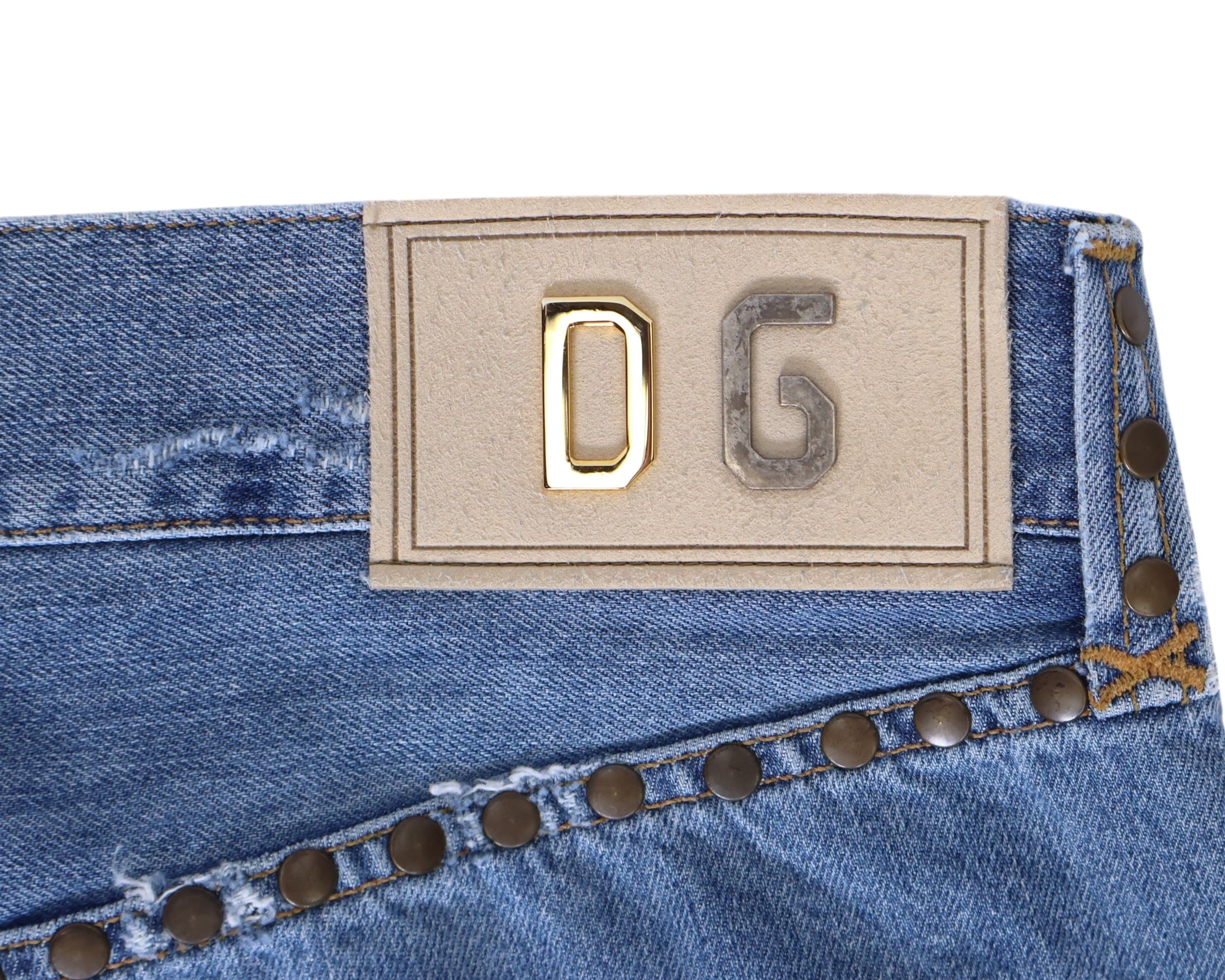 Close-up of vintage Dolce & Gabbana light-wash blue denim jeans/denim trousers showing a beige DG logo patch with mixed-metal letters, studded rivet trim and contrast stitching. High-end designer detail shot—rare, unique, luxury denim.