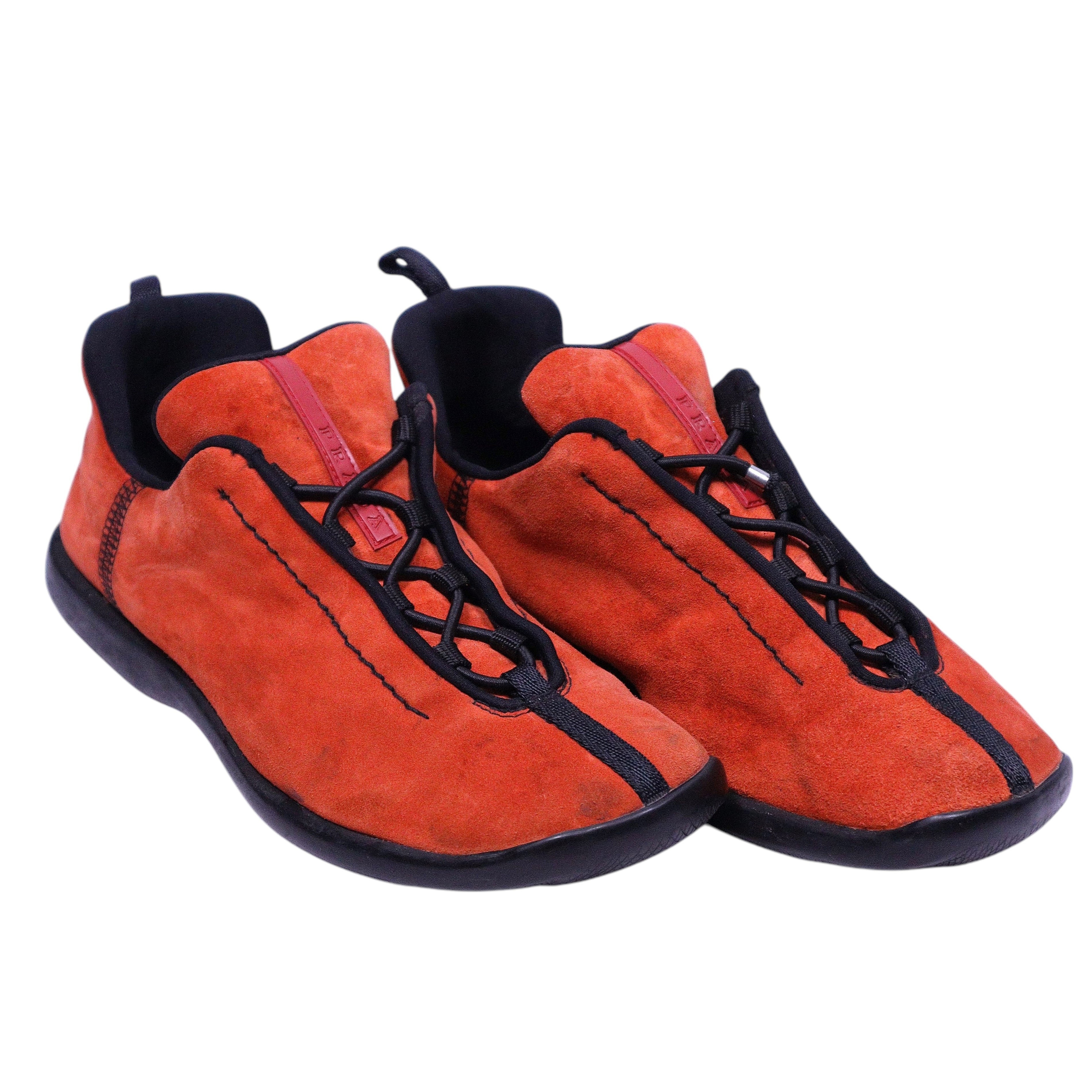 A pair of burnt orange suede trainer shoes with black accents and laces. Prada burnt-orange suede low-top trainers with contrast black piping, speed-lace front, soft textile collar and rounded toe on a rubber sole; vintage, luxury, rare.