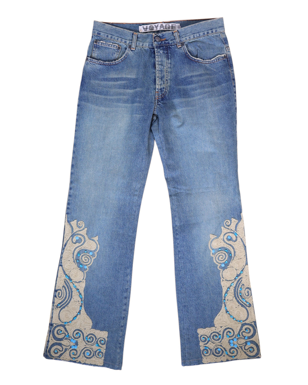 A pair of blue jeans with stone embroidery on the lower legs. Medium-blue denim jeans with straight legs and ornate cream appliqué at the hems, traced with turquoise stones and swirl embroidery. Five-pocket, belt-loop waist and zip fly; vintage/designer, rare statement pair.