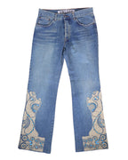 A pair of blue jeans with stone embroidery on the lower legs. Medium-blue denim jeans with straight legs and ornate cream appliqué at the hems, traced with turquoise stones and swirl embroidery. Five-pocket, belt-loop waist and zip fly; vintage/designer, rare statement pair.
