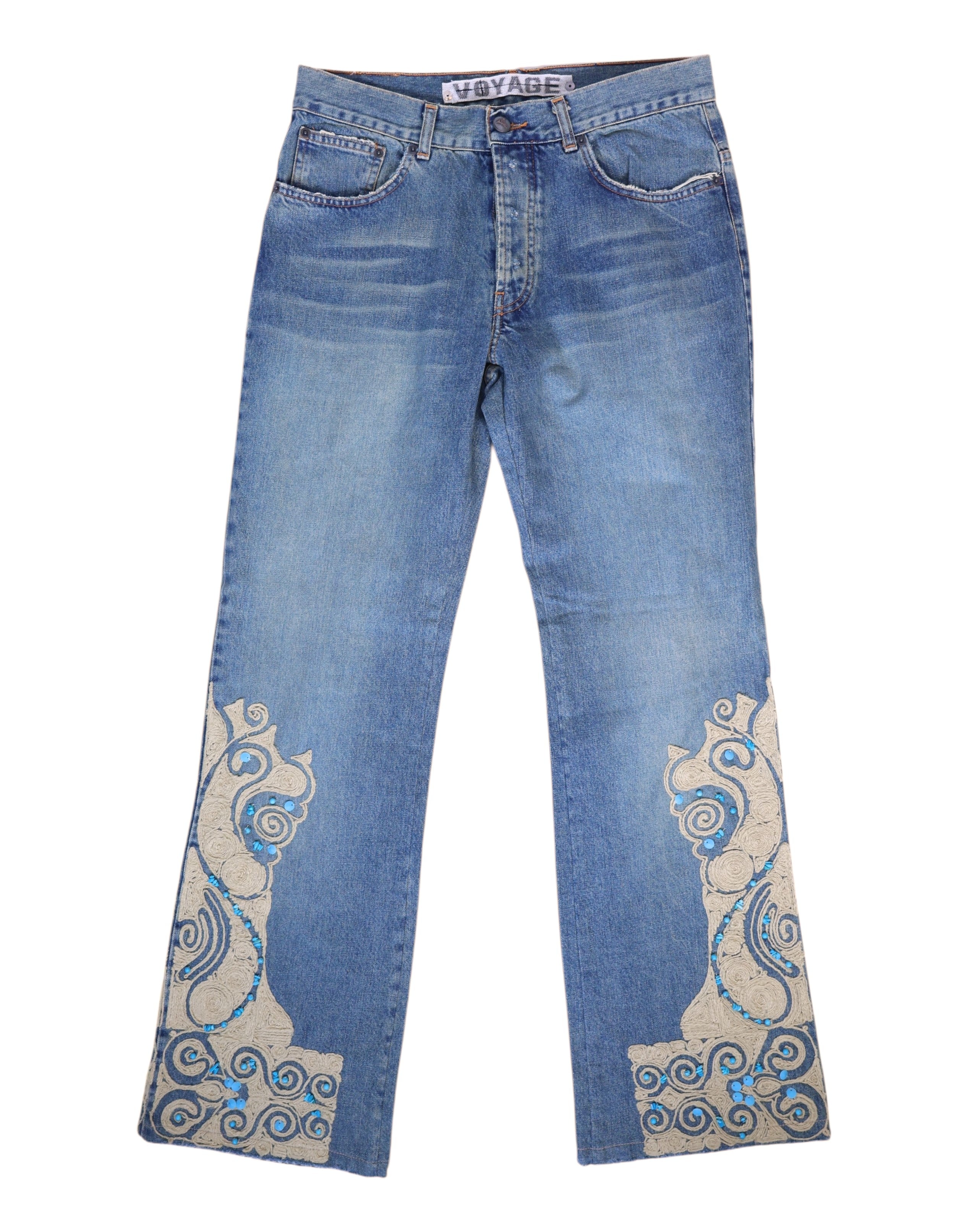 A pair of blue jeans with stone embroidery on the lower legs. Medium-blue denim jeans with straight legs and ornate cream appliqué at the hems, traced with turquoise stones and swirl embroidery. Five-pocket, belt-loop waist and zip fly; vintage/designer, rare statement pair.
