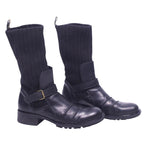 A pair of black leather boots with ribbed knit cuffs and buckle details, set against a white background. Roberto Cavalli black leather biker boots with ribbed knit shafts, buckle straps, round toes, and lug soles.
