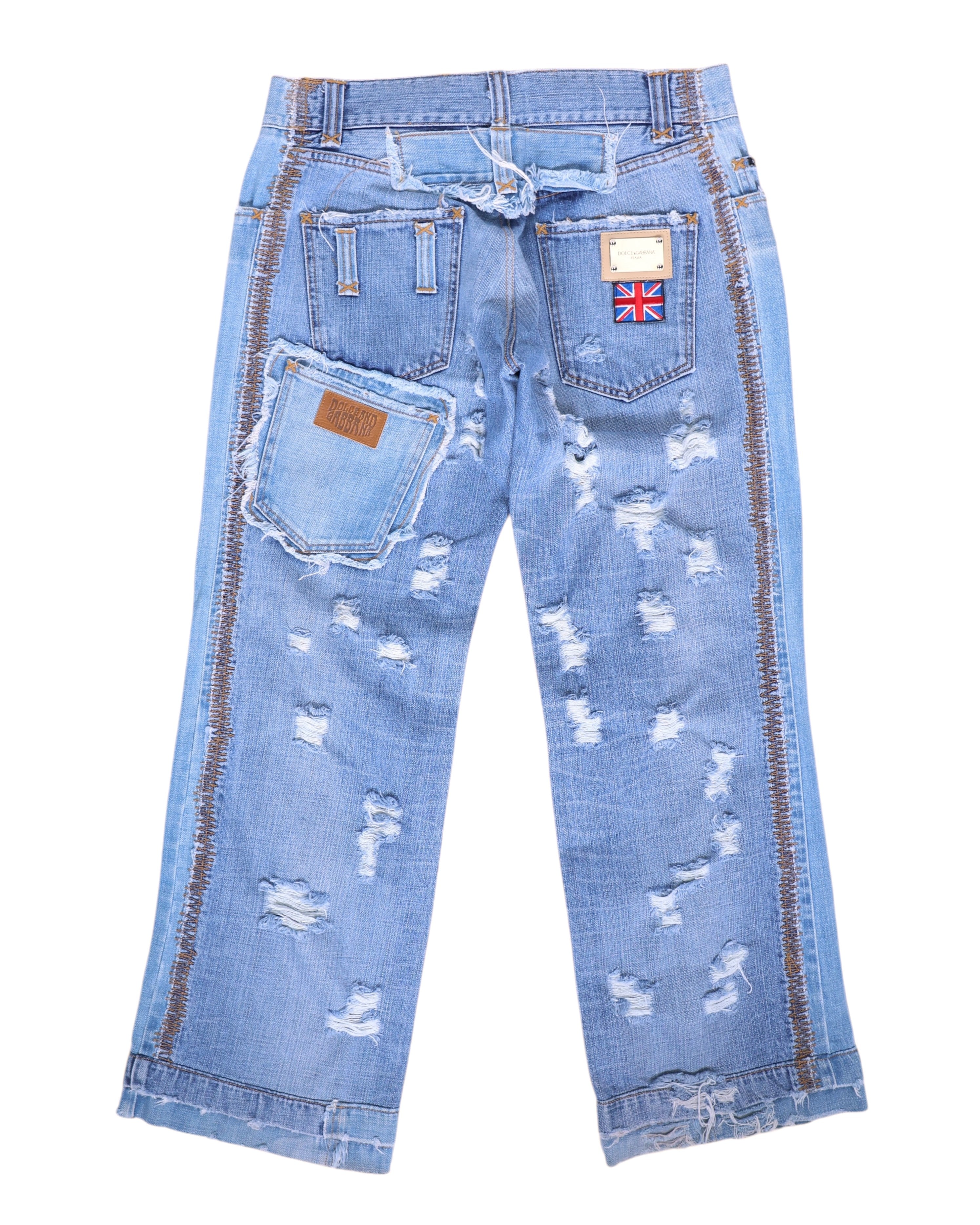 Vintage Dolce & Gabbana Silver Label light-wash denim jeans/denim trousers, back view, heavily distressed with shredded rips, raw frayed hems and a straight, slightly cropped leg. Contrast side stitching, multiple back pockets including an angled oversized pocket with leather logo tab, plus a Union Jack patch—unique luxury designer piece laid flat on white.