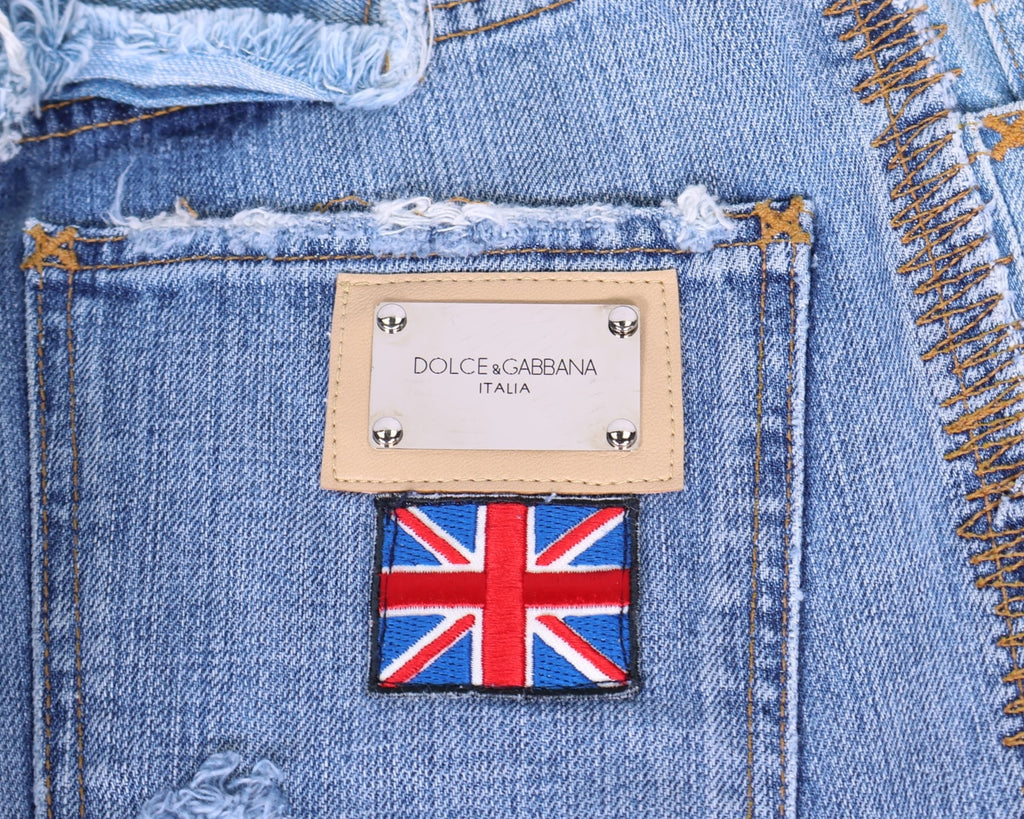 Close-up of vintage Dolce & Gabbana light-wash denim, showing a metal logo plaque on a tan leather frame above an embroidered Union Jack patch, with heavy distressing, frayed edges and contrast whipstitch. Luxury designer detail shot.