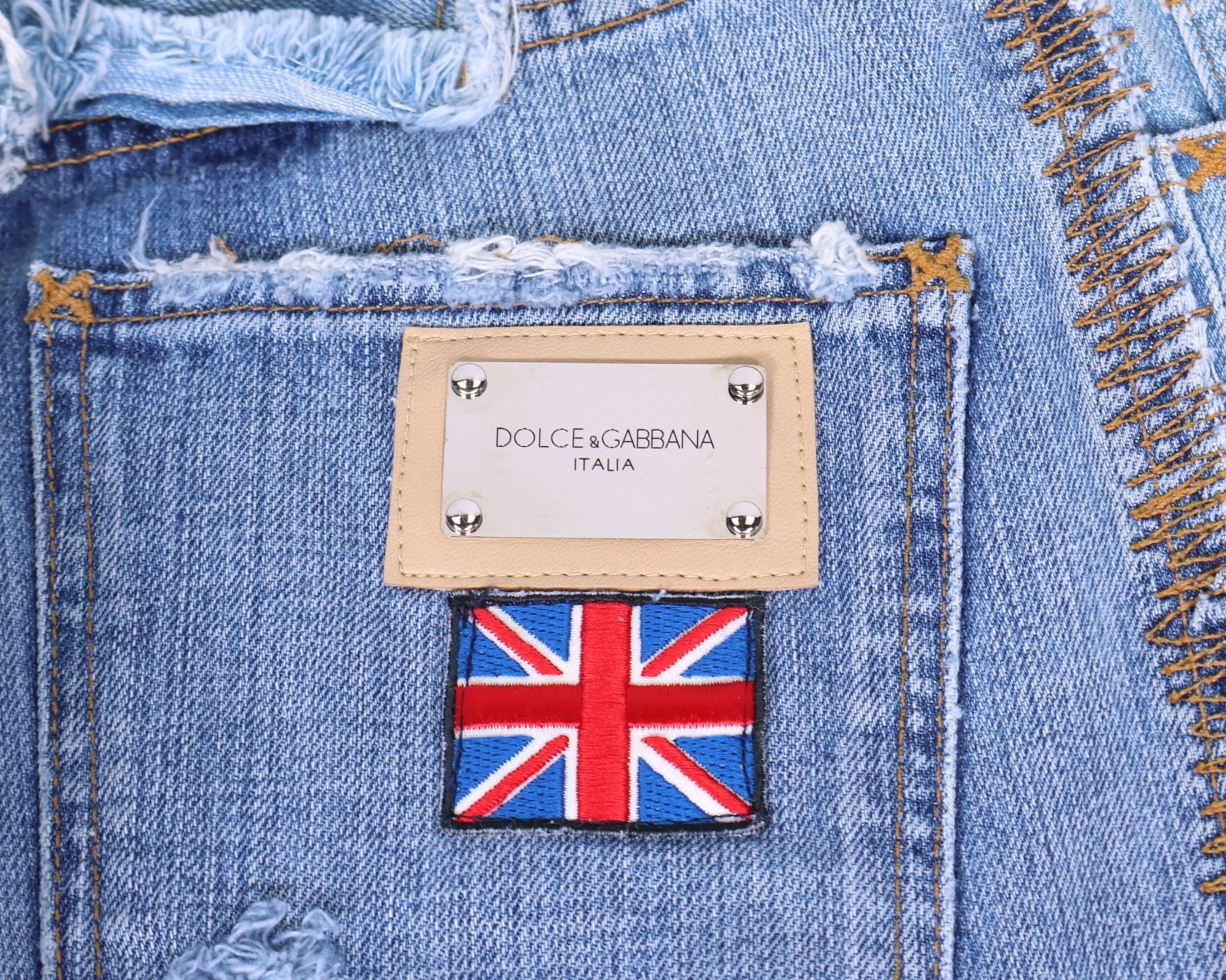 Close-up of vintage Dolce & Gabbana light-wash denim, showing a metal logo plaque on a tan leather frame above an embroidered Union Jack patch, with heavy distressing, frayed edges and contrast whipstitch. Luxury designer detail shot.