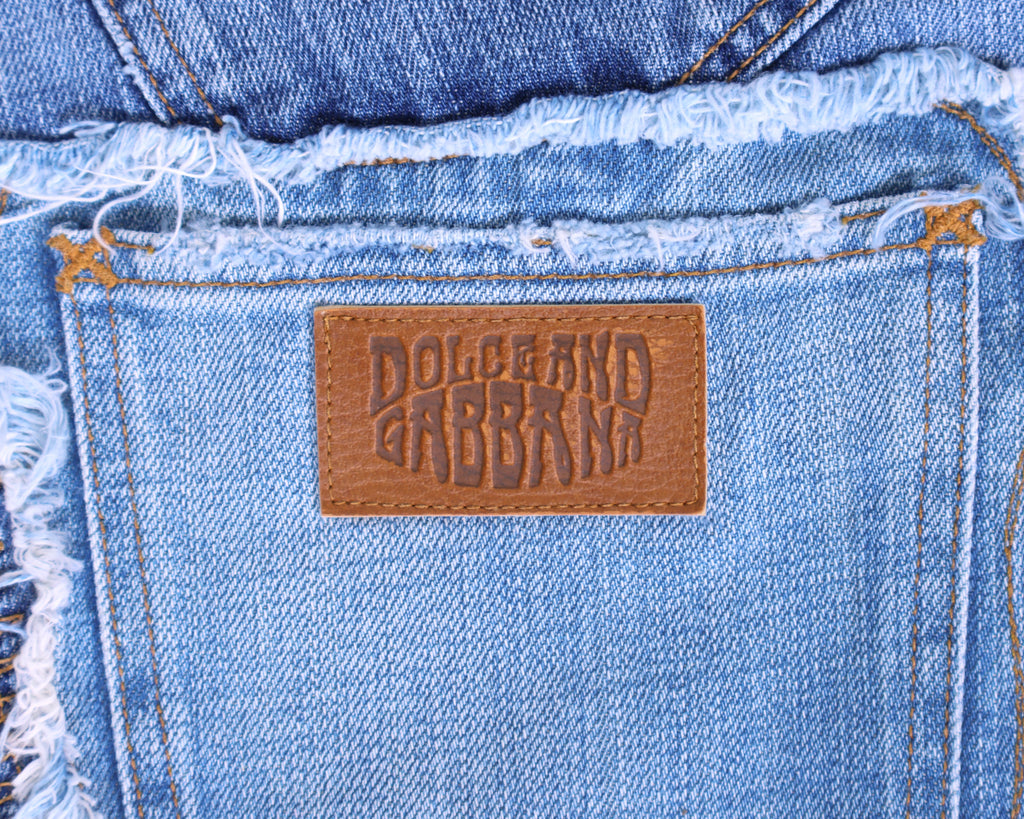Detail shot of vintage Dolce & Gabbana light-wash denim back pocket with an embossed tan leather logo patch and raw, frayed waistband stitching. High-end designer jeans/denim, worn-in and rare.