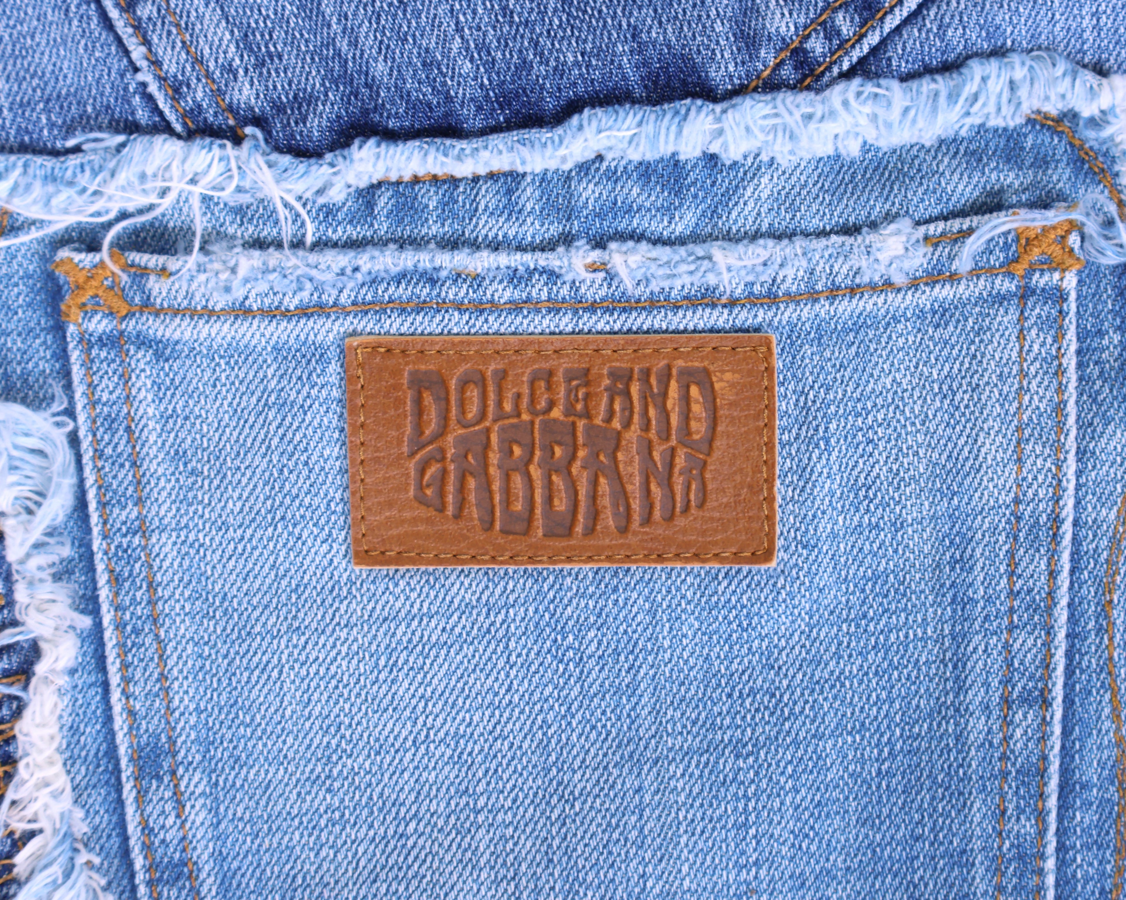 Detail shot of vintage Dolce & Gabbana light-wash denim back pocket with an embossed tan leather logo patch and raw, frayed waistband stitching. High-end designer jeans/denim, worn-in and rare.