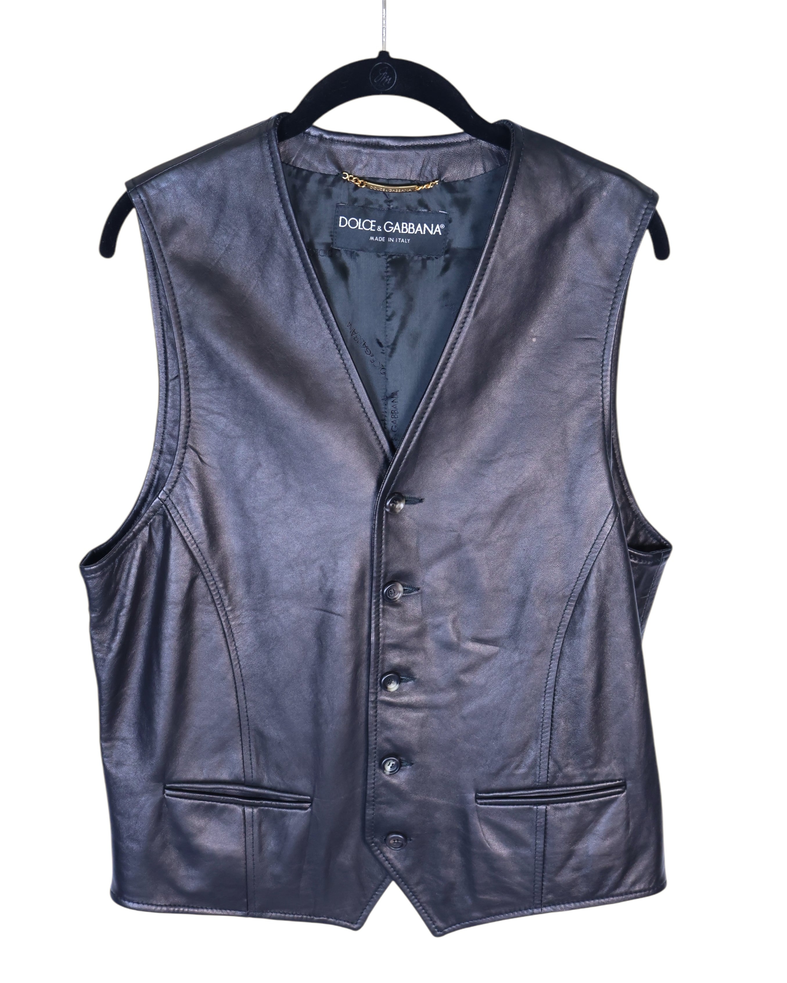 Dolce & Gabbana black leather vest with buttons, collarless, sleeveless, displayed on a hanger. Dolce & Gabbana (D&G) black leather vest / waistcoat / gilet—Made in Italy, genuine smooth calf leather, V-neck with six-button front, tailored darts, welt pockets, satin lining; sleek smart-casual to formal layer with rock-chic edge, Y2K runway vibe; Italian luxury craftsmanship, unisex styling over shirts, tees or under blazers.