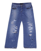 A pair of blue denim jeans with a distressed pattern and charms attached to the pockets. Medium-blue distressed denim jeans with heavy rips and frayed patches, straight-leg five-pocket cut with belt loops and zip fly. Black dome studs line the front pockets with scattered metal charms; vintage/designer, rare statement pair.