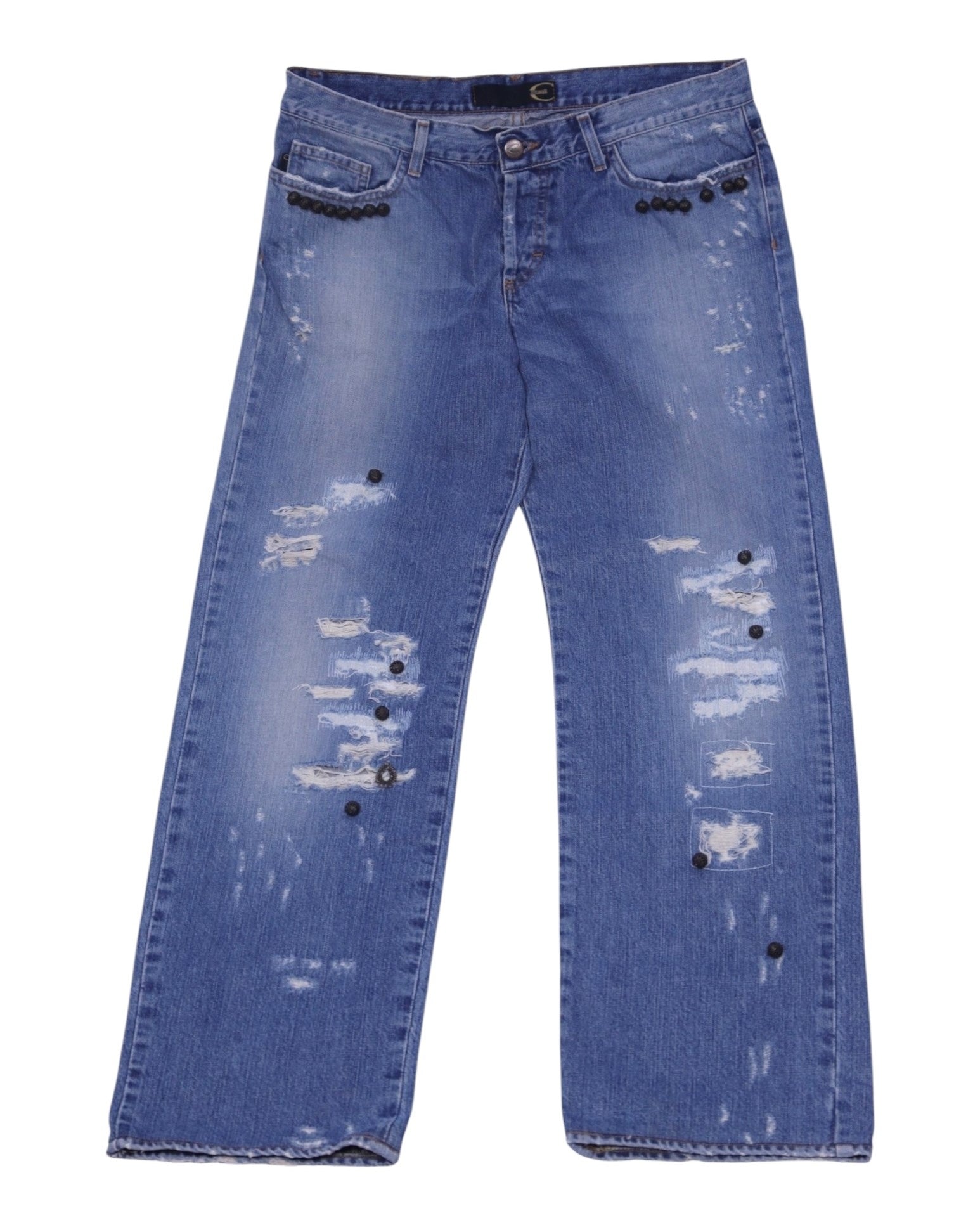 A pair of blue denim jeans with a distressed pattern and charms attached to the pockets. Medium-blue distressed denim jeans with heavy rips and frayed patches, straight-leg five-pocket cut with belt loops and zip fly. Black dome studs line the front pockets with scattered metal charms; vintage/designer, rare statement pair.