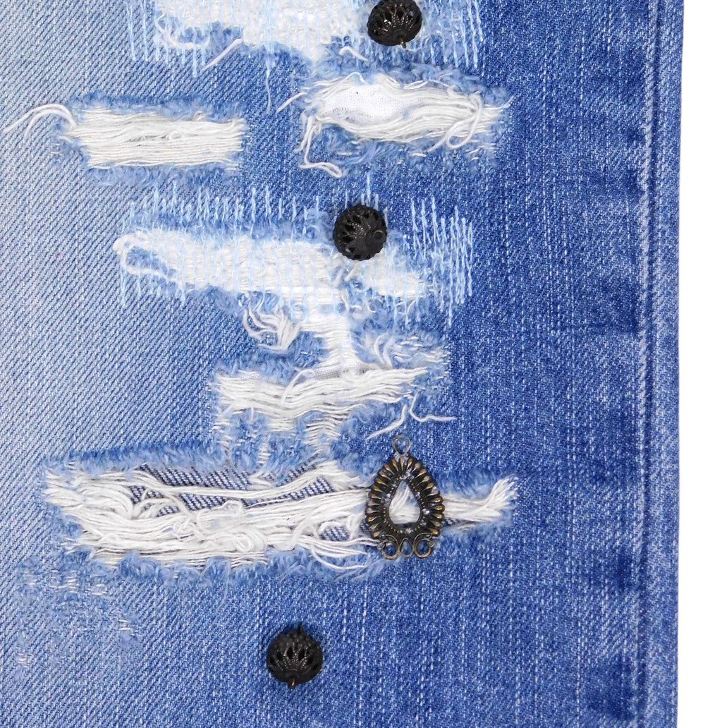 Close-up of distressed blue denim showing shredded rips, white threads and black metal studs with an antique teardrop charm. Textured, punk-leaning detail from a vintage designer jean.