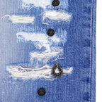 Close-up of distressed blue denim showing shredded rips, white threads and black metal studs with an antique teardrop charm. Textured, punk-leaning detail from a vintage designer jean.