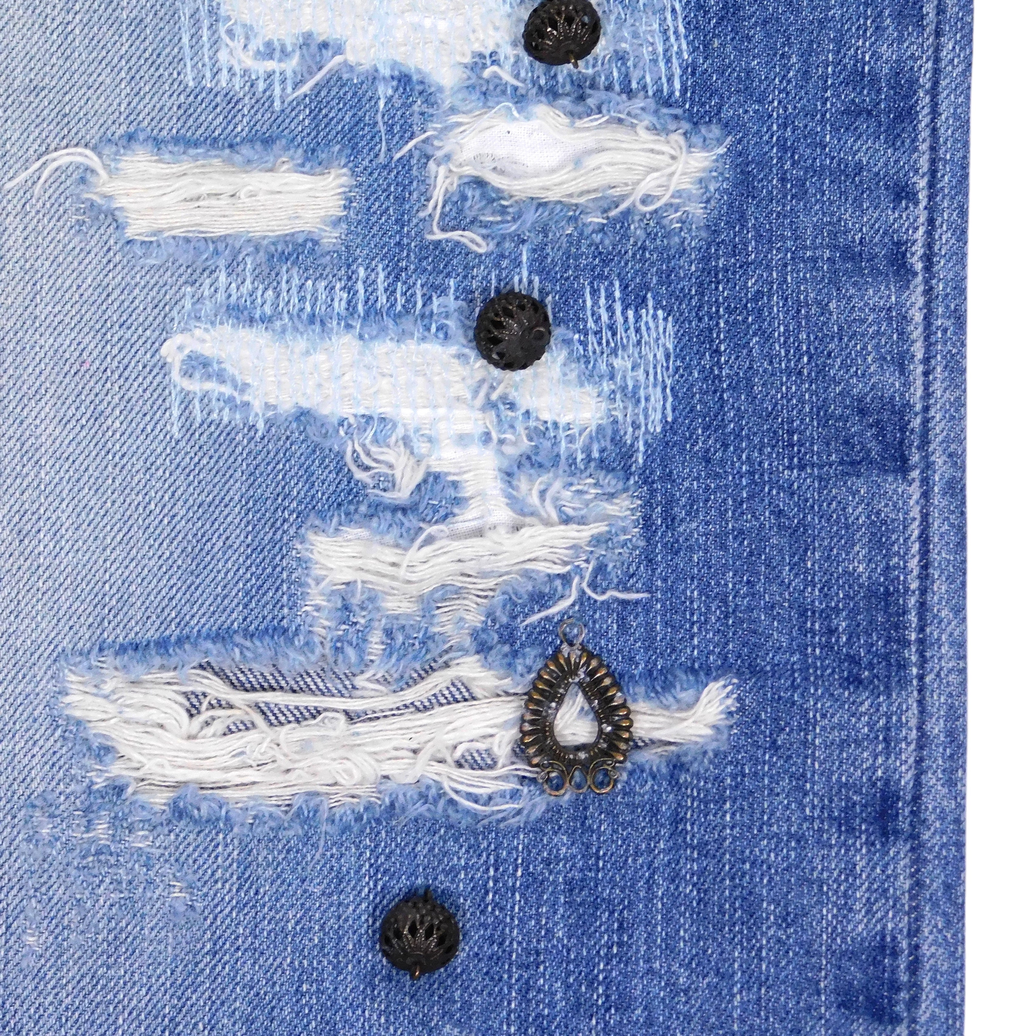 Close-up of distressed blue denim showing shredded rips, white threads and black metal studs with an antique teardrop charm. Textured, punk-leaning detail from a vintage designer jean.